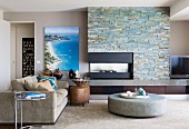 Runder, gepolsterter Couchtisch und Sofa vor Natursteinwand mit offenem Kamin in modernem Ambiente