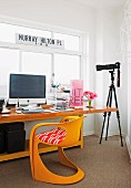 Schalenstuhl vor Schreibtisch mit Computer; daneben Kamera auf Stativ vor Fenster