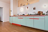Long counter with red accents in open-plan kitchen with oak floor and aperture in wall above wall lamps
