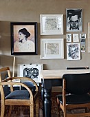 Rustikale Holzstühle mit schwarzem Bezug auf Sitzfläche und Holztisch mit schwarz lackierten, gedrechselten Beinen, vor hellgrau getönter Wand mit gerahmten Bildern