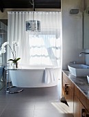 Modern bathroom - washstand with wooden base cabinet built into niche, free-standing bathtub in front of closed, translucent curtains at window