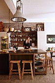 Woman standing behind counter in rustic, country-house-style kitchen with industrial lamps and ceramic crockery on open-fronted shelving
