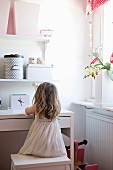 Little girl sitting on stool at white desk and storage boxes on floating shelves