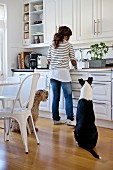 Woman at sink in fitted kitchen, two dogs sitting on floor and dining area with vintage chairs