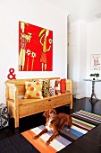 Dog lying on striped rug in front of rustic bench with scatter cushions below artwork