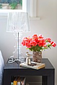 Bouquet of red flowers in vase next to transparent, plastic table lamp on dark bedside table