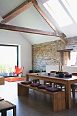 Esstisch und Bank aus Holz mit Kissen in renoviertem, offenem Wohnraum mit sichtbarer Dachkonstruktion aus Holz