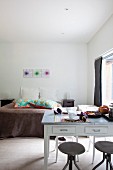 Drehhocker vor Küchentisch mit Frühstücksgedecken, im Hintergrund Doppelbett in minimalistischem Schlafzimmer