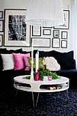Round, modern coffee table on dark, long-pile rug in front of black sofa with row of scatter cushions below gallery of pictures on wall