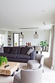 Graue Polstersessel im Fiftiesstil und Sofa um Bodentisch aus rustikalen Vierkanthölzern in minimalistischem Wohnraum, im Hintergrund skandinavische Designerleuchten über Essplatz