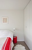 Spherical sconce lamp above red, cylindrical bedside cabinet between bed with red and white blanket and white fitted wardrobes