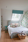 Bett mit gepolstertem Kopfteil vor Dachflächenfenster in Kinderzimmer, Wäschekorb mit Stofftieren an Bettende