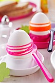 Boiled eggs wrapped in brightly coloured woollen yarn in pink and white eggcups with plastic spoons