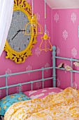 Child's bed with retro, turquoise metal frame in corner below large wall clock with yellow frame on pink wallpaper with ornamental pattern