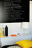White table lamp and colourful candlesticks on sideboard below black panel with white lettering on wall