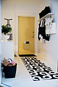 Black and white patterned runner on white hallway floor, wall-mounted coat rack and front door painted pale yellow with letterbox cage