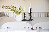 Porzellankrug mit Blätterzweig und Kerzenständer auf Tisch, vorne teilweise sichtbare, geschwungene Rückenlehne eines Küchenstuhls
