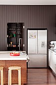Küchenblock mit Holzhockern, dahinter ein schwarzer Schrank mit Falttür und ein Kühlschrank als Einbauten in grauer Wandvertäfelung