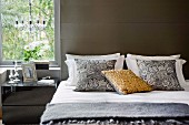Kissenstapel auf modernem Doppelbett vor grauer Wand, seitlich verspiegeltes Nachtkästchen am Fenster