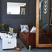 Seating area with brown leather sofa, white trunk table, potted plants and mirror with frame painted the same slate grey as wall; open glass door to one side