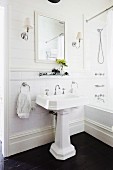 Vintage pedestal sink on dark wooden floor, framed mirror and symmetrical lamps on white wooden wall in traditional bathroom
