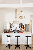 Black metal bar stools made from old tractor seats at white kitchen counter below antique chandelier in modern interior with woman in background