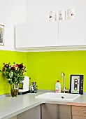 Kitchen counter with lime green splashback, white wall units, bouquet, coffee machine and cassette-tape chopping board