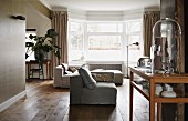 Wohnraum in warmen Grau- und Holztönen mit Polsterelementen vor einem Erkerfenster; Vitrinentisch im Vordergrund