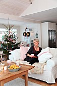 Woman sitting on white sofa next to decorated Christmas tree in rustic living room