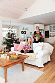 Familie auf und hinter dem Sofa neben geschmücktem Weihnachtsbaum in offenem, ländlichem Wohnzimmer