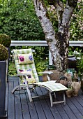 Rustic wooden lounger with cushions next to urns and tree on dark grey wooden deck