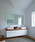Floating, designer washstand with two countertop basins, wooden counter and white base unit with drawers below custom mirror in minimalist bathroom
