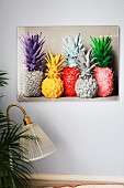 Abbildung quietschbunt lackierter Ananasfrüchte an lichtgrauer Wand, Vintage-Leuchte und Palmenblätter im Vordergrund