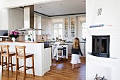 Girl in open-plan, white country-house kitchen with wooden bar stools at breakfast bar integrated into kitchen counter