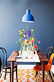 Bottles of spring flowers on simple wooden table with white runner, Thonet chairs and classic shell chair against blue-painted wall