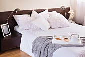 Frühstück im Bett, weiße Kissen teilweise mit Rüschen-Stoff auf Doppelbett, seitlich Retro-Tischleuchte auf Nachtkästchen an halbhoher, dunkler Holzwand