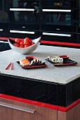 Detail of island counter with black and red accents, pale stone surface, bowl of tomatoes and two plates of sushi
