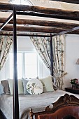 Bett mit Himmelgestell aus dunklem Holz, Kissenstapel, vor Fenster in Schlafzimmer mit Holzbalkendecke