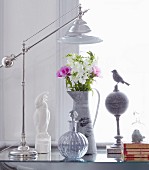 Table lamp with shiny chrome frame, spherical bottle with stopper and bouquet in zinc jug on glass table top