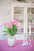 Vase of pink tulips and white candles in candlesticks on pink and white patterned tablecloth