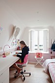 Girl sitting on pink swivel chair at desk worksurface along one wall of attic bedroom