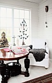 Runder Holztisch mit gedrechselten Beinen, schwarz lackiert und weisser Polstersessel vor Sprossenfenster in holzverkleideter Zimmerecke