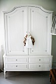 Antique, white-painted wardrobe on delicate feet with small fabric bag and wreath hanging from key