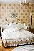 Doppelbett mit weißem, geschnitztem Kopfteil, vor Wand mit Tapete, in traditionellem Schlafzimmer mit weissen und hellbraunen Farbtönen