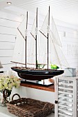 Splendid model of sailing boat on half-height wall in Scandinavian interior; wicker tray and vintage lantern in foreground