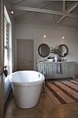 Free-standing bathtub below window and long washstand with base cabinet in spacious bathroom with rustic ambiance