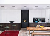 Rustic wooden table and bench in front of open-plan kitchen, delicate, retro-style bar stools at monolithic counter in front of white fitted cupboards