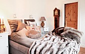 Comfortable, upholstered bed with soft fur blankets and several beige scatter cushions; antique longcase clock against wall