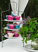 Muffins in colourful paper cases on cake stand and vase of lilac on garden table