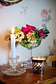 Lit candle in vintage, glass candlestick next to tealight holder in front of dish of flowers